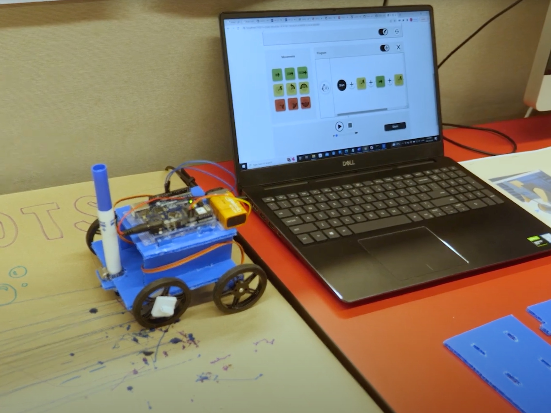 Image of a blue robot with four black wheels and a blue marker mounted on the front of the chassis. Next to it, a black laptop computer on an orange desk is running Weavly in a web browser.