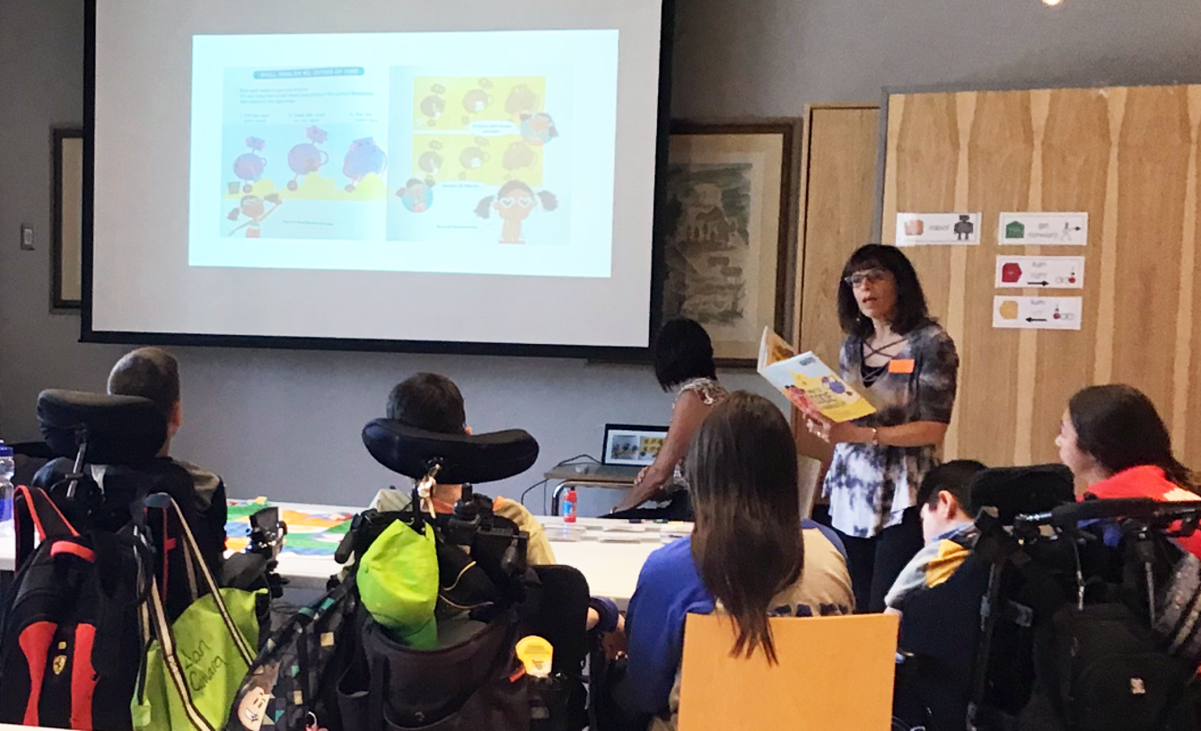 An instructor is reading from a picture book that is also projected on the screen for a group of students sitting on wheelchairs. 